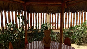 Una mesa de madera con una estatua en el centro, rodeada de sillas, bajo una estructura de techo de paja con pilares de bambú.