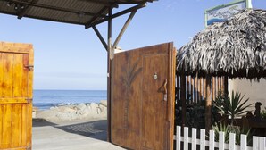 Un portón de madera con diseño de palmera que da paso a una zona de playa con una estructura de techo de paja y un letrero que dice "The Cottages by the Sea".