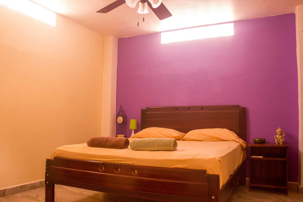 Une chambre à coucher avec un lit en bois, des murs violets, un ventilateur de plafond, une table de chevet avec une horloge et une petite statue.