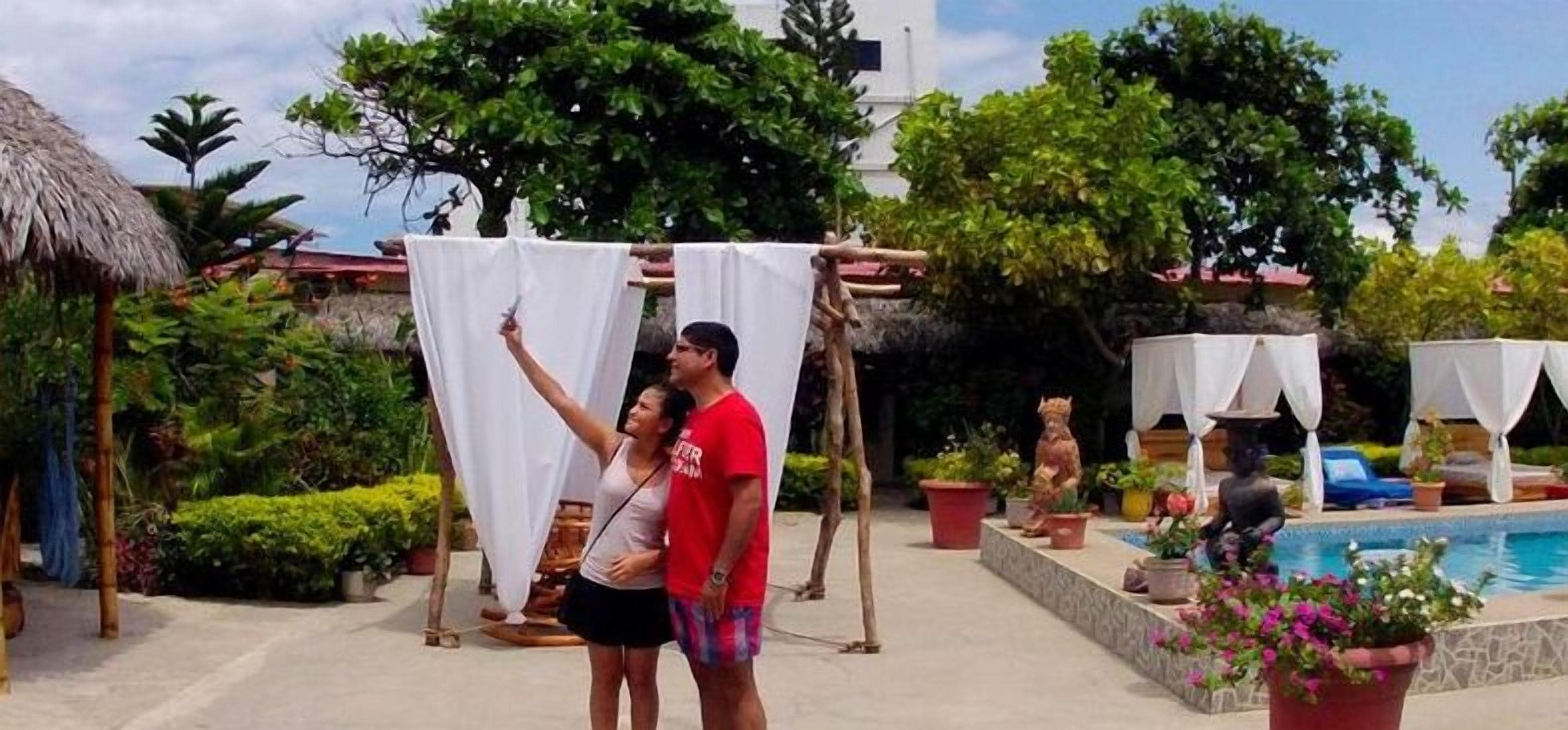 Una pareja bajo un toldo cerca de una piscina, rodeada de vegetación exuberante y plantas en macetas.