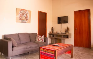 Una sala de estar con un sofá gris, una mesa de centro colorida, un televisor de pantalla plana montado en la pared y dos puertas de madera.