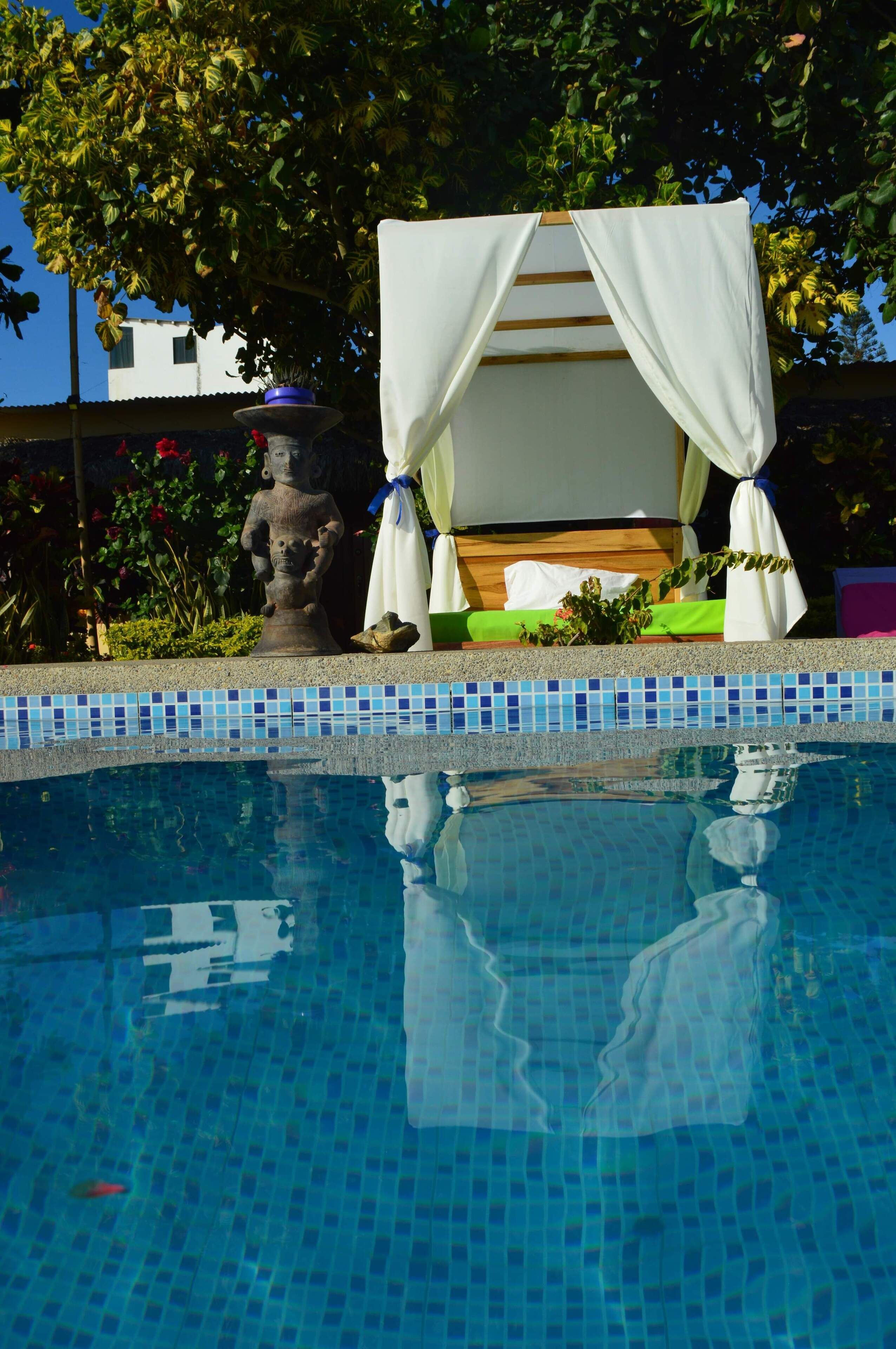 A pool with a tiled edge, a statue, and a covered seating area with white curtains.