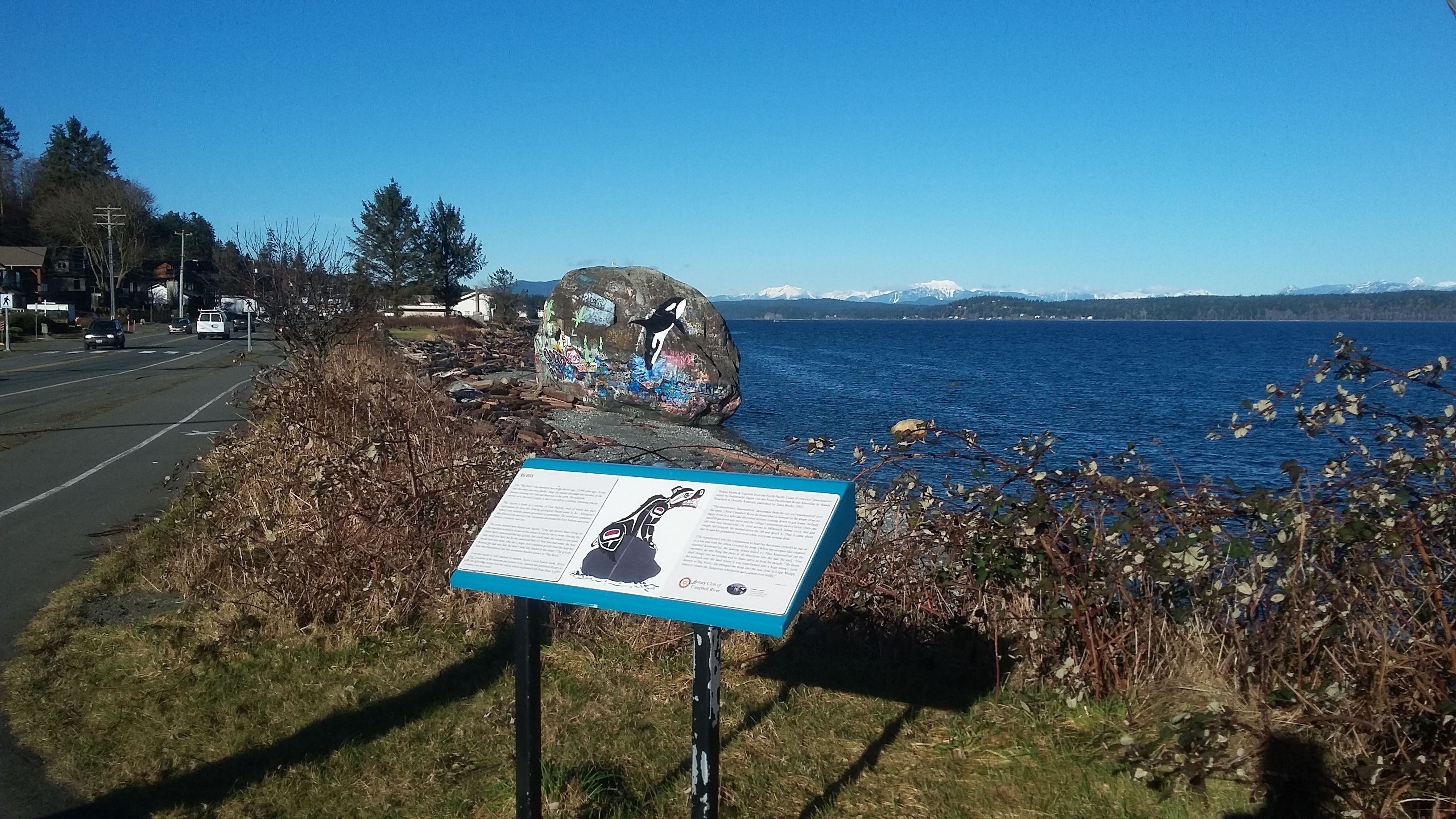 Un cartel a la carretera con un mapa y texto, una vía con vehículos, y una masa de agua con montañas al fondo.