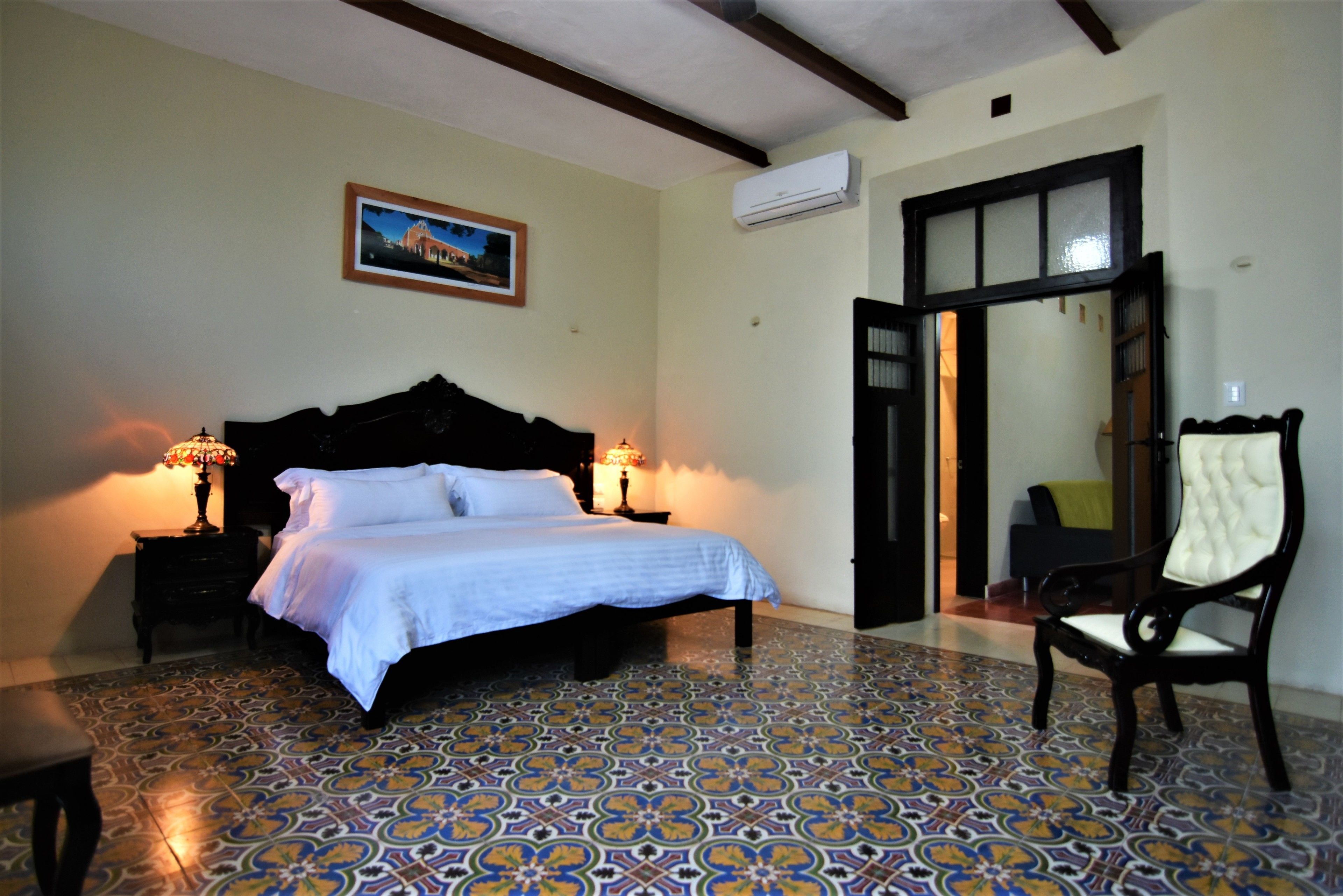 Une chambre à coucher comprenant un lit, deux tables de chevet avec des lampes, une chaise, une porte et un climatiseur fixé au mur.