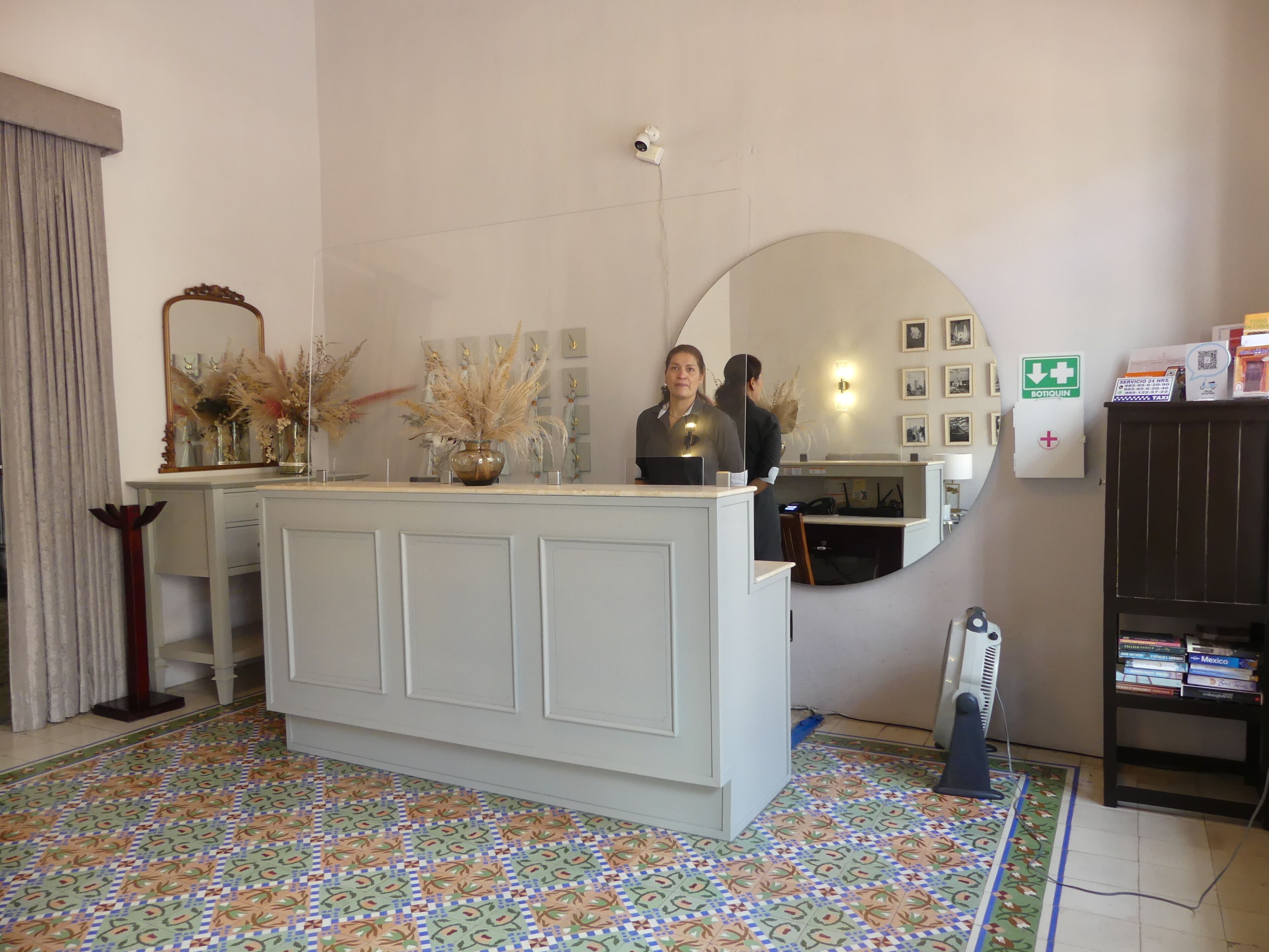 A reception area with a counter, a mirror, a vase with dried plants, a wall-mounted security camera, framed pictures, a sign indicating a pharmacy, and a bookshelf with books.