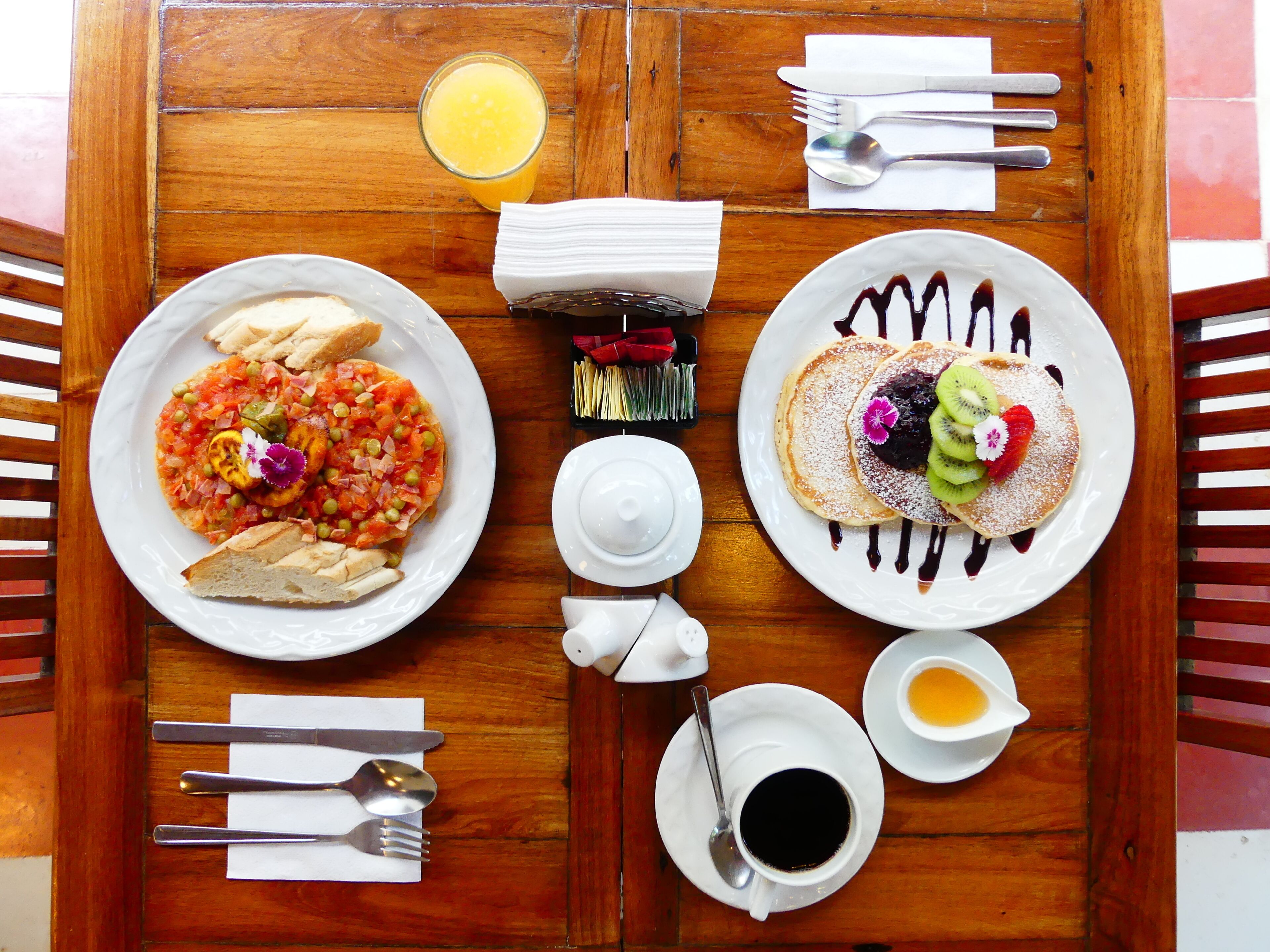 Una mesa de madera con platos de desayuno, cubiertos y un vaso de zumo de naranja.