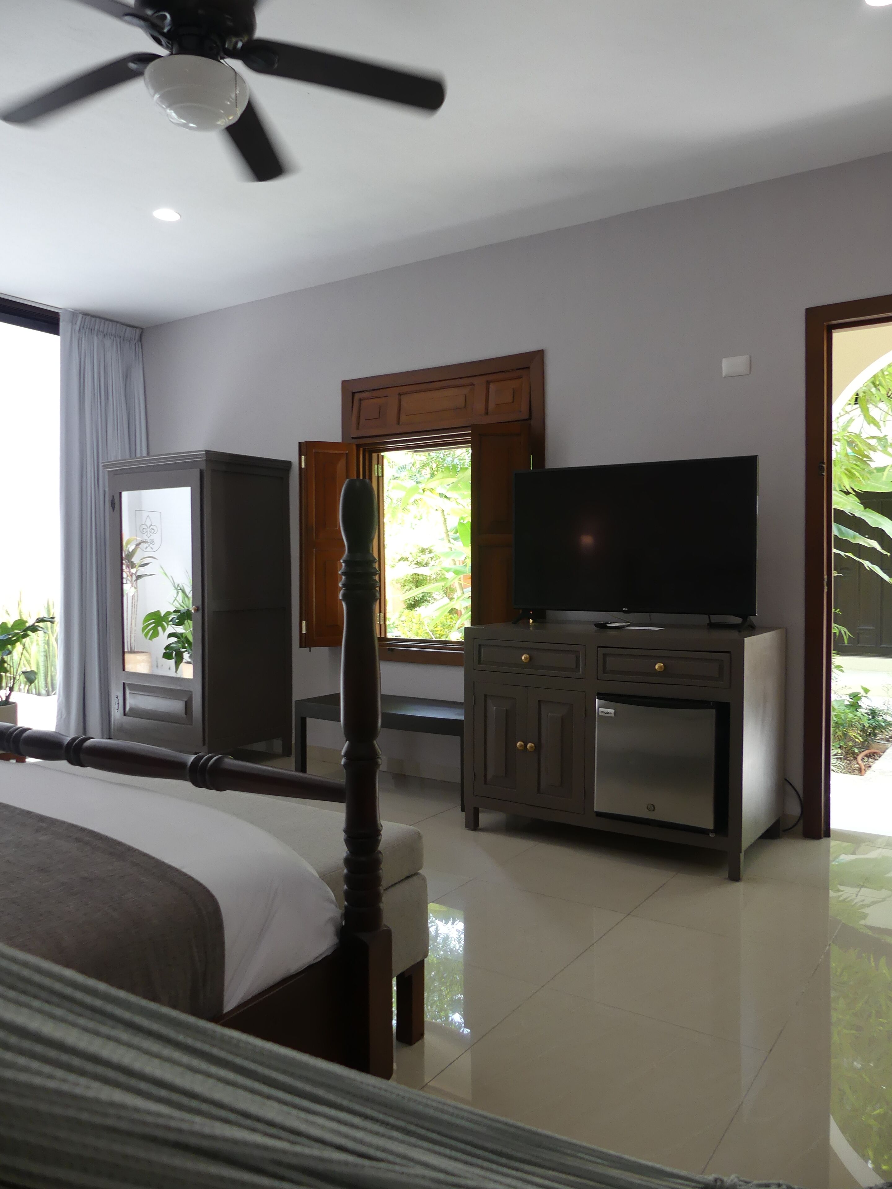Un dormitorio con cama, televisor sobre un mueble, ventilador de techo y ventana con vista a follaje.