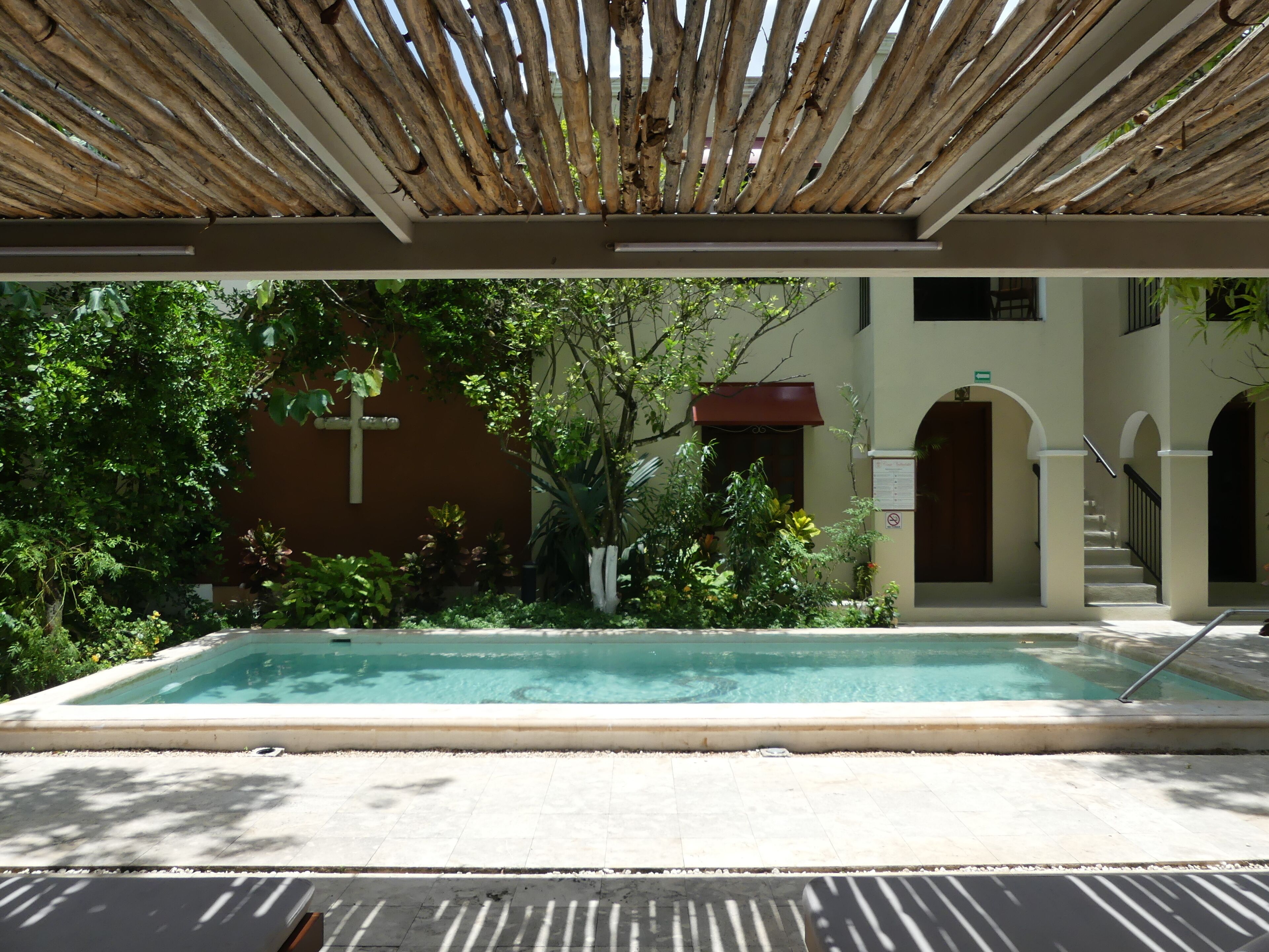 An outdoor swimming pool with a wooden pergola overhead, surrounded by lush greenery and a building with a red awning and white columns.