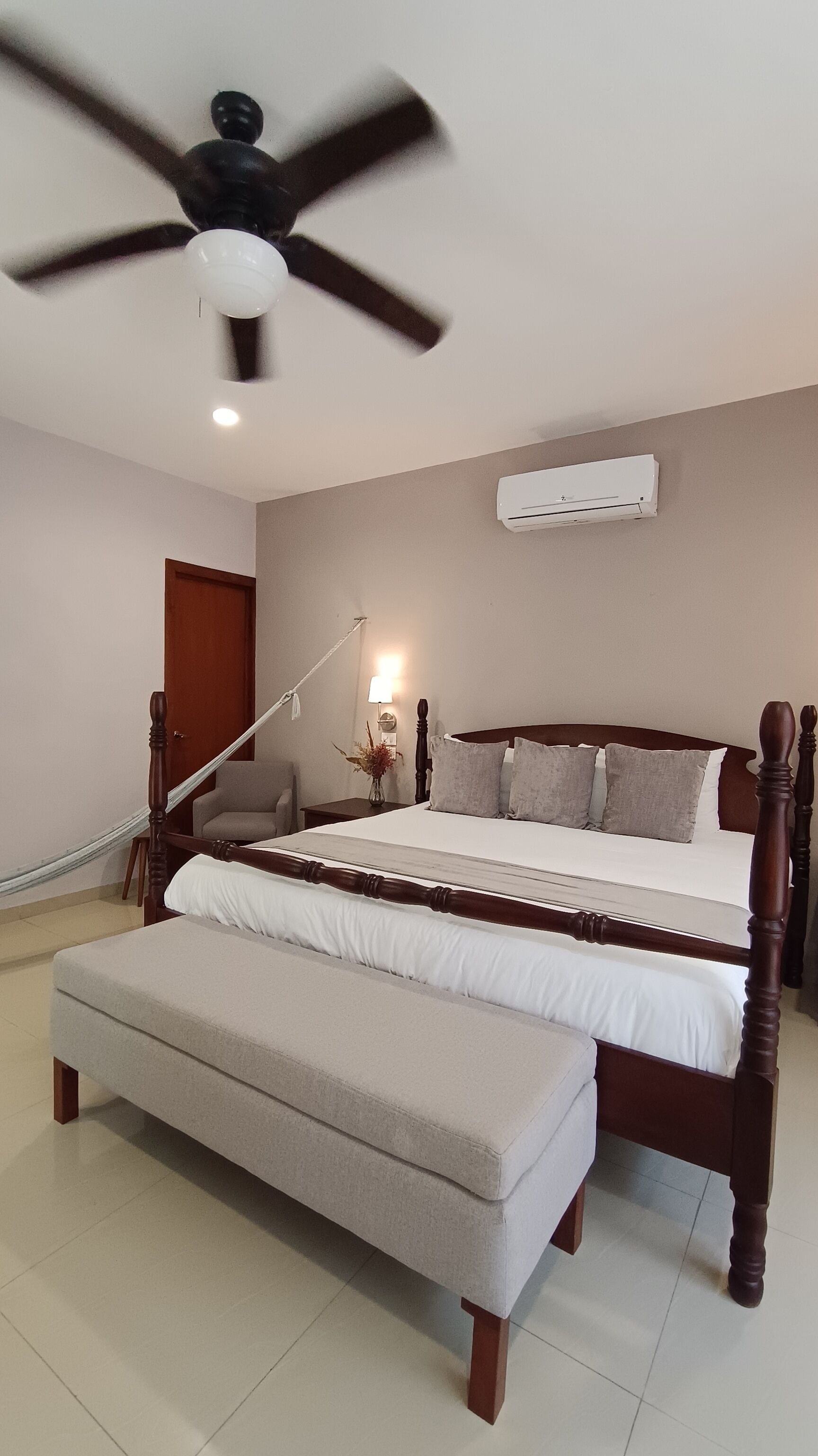 Un dormitorio con cama, un banco, ventilador de techo, equipo de aire acondicionado, lámpara y una puerta.