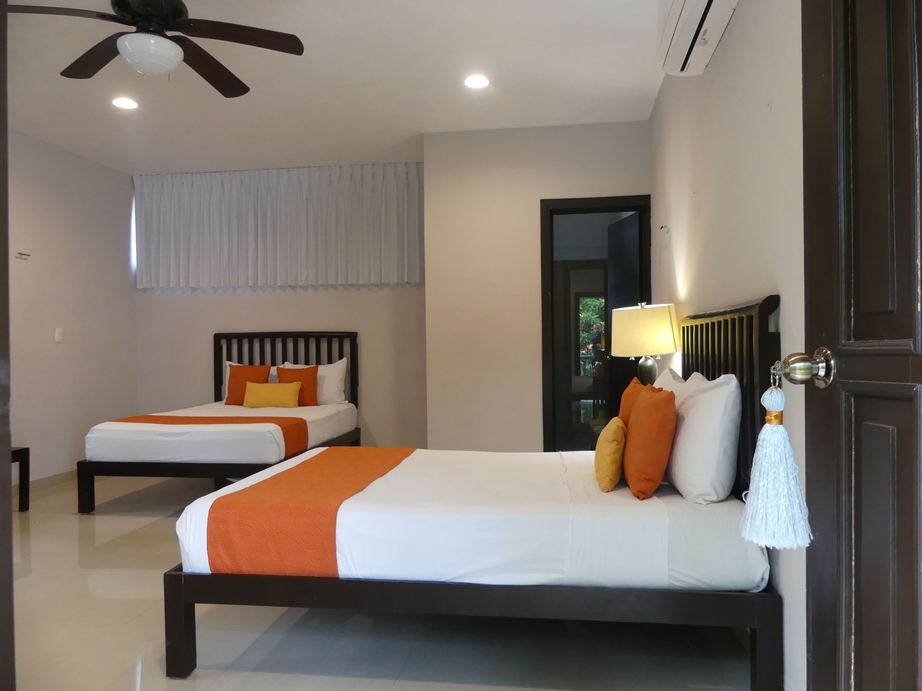 A hotel room with two beds, a ceiling fan, a window with curtains, and a door with a mirror.