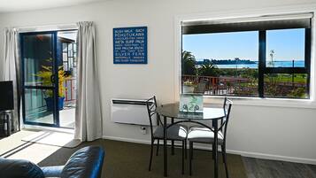 A room with a dining table, chairs, a sofa, a large window with a view of the sea, and a sign listing amenities.