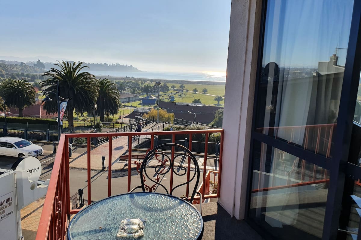 Ein Balkon mit Glastisch und Stühlen, der auf eine Küstenlandschaft mit Palmen und freiem Blick auf das Meer blickt.