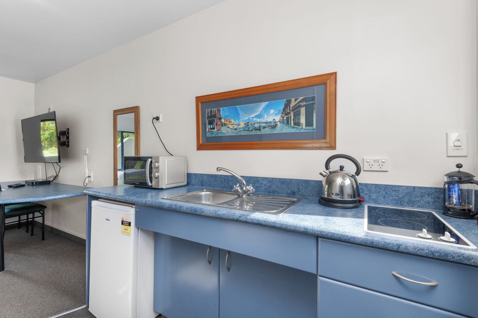 Een praktische keuken met een spoelbak, waterkoker en een kleine koelkast. Een flatscreen televisie is aan de muur bevestigd, en er hangt een lijst boven de aanrecht.