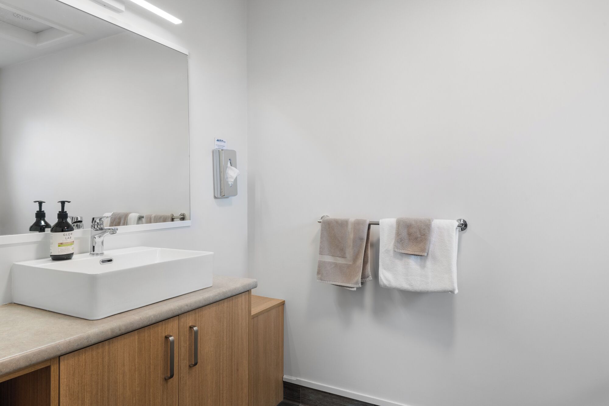 A modern bathroom with a white sink, wooden cabinet, and two towels on a rack.