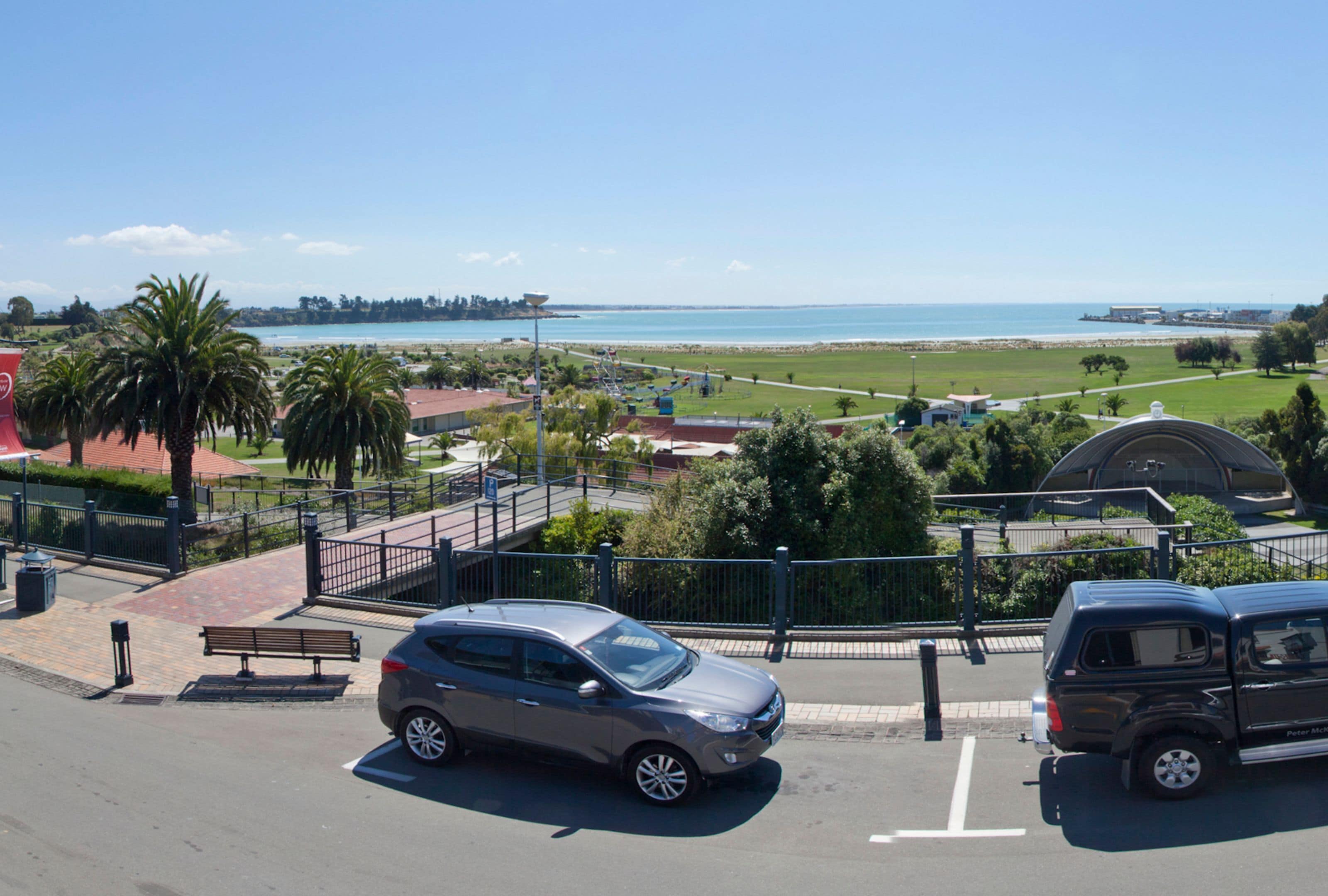 Un estacionamiento con coches y un banco, un sendero y vistas al mar y al verdor.