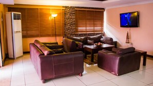 A living room with a sofa set, a coffee table, a flat-screen TV mounted on the wall, and a window with blinds. - EED Pension Boutique Hotel (Lagos)