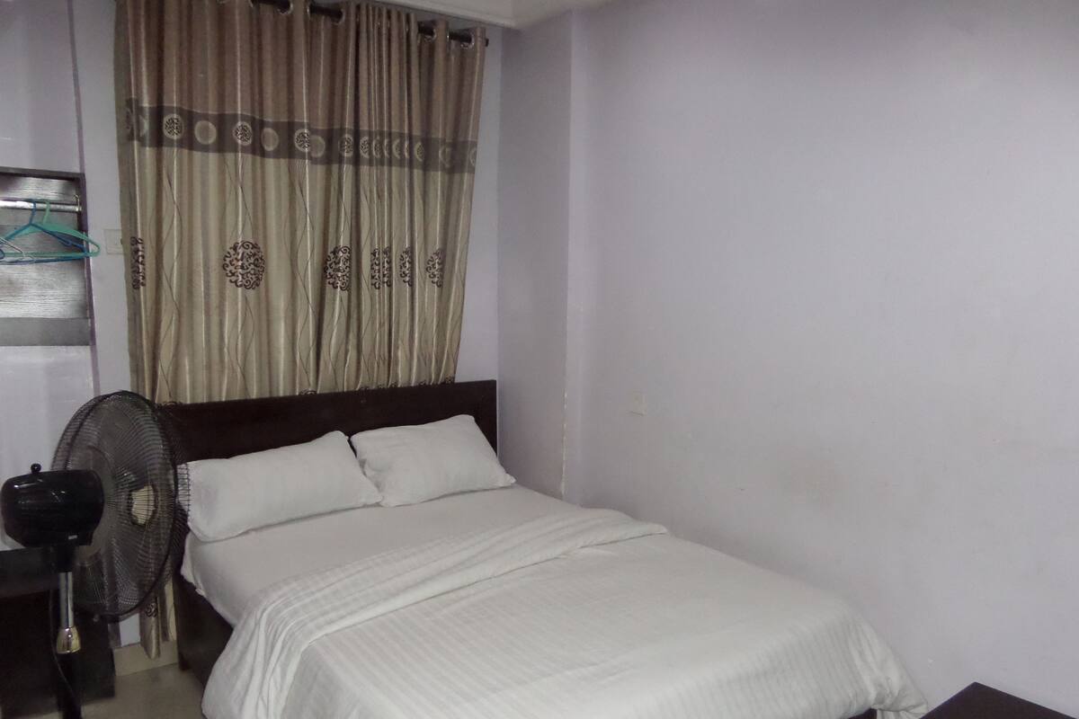 A single bed with white bedding, a dark headboard, a standing fan, and a curtain with a patterned design.