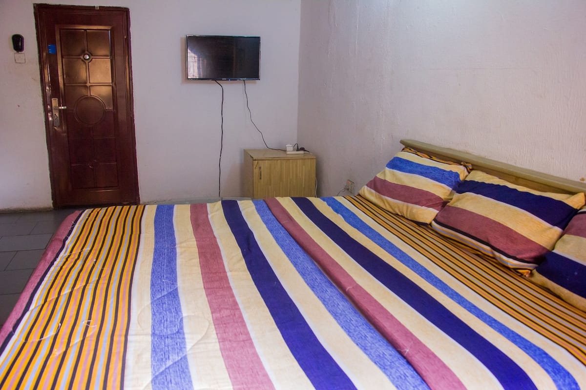 Ein Schlafzimmer mit gestreifter Bettdecke, einem Holz-Kopfteil, einem an der Wand montierten Fernseher und einem kleinen Nachttisch.