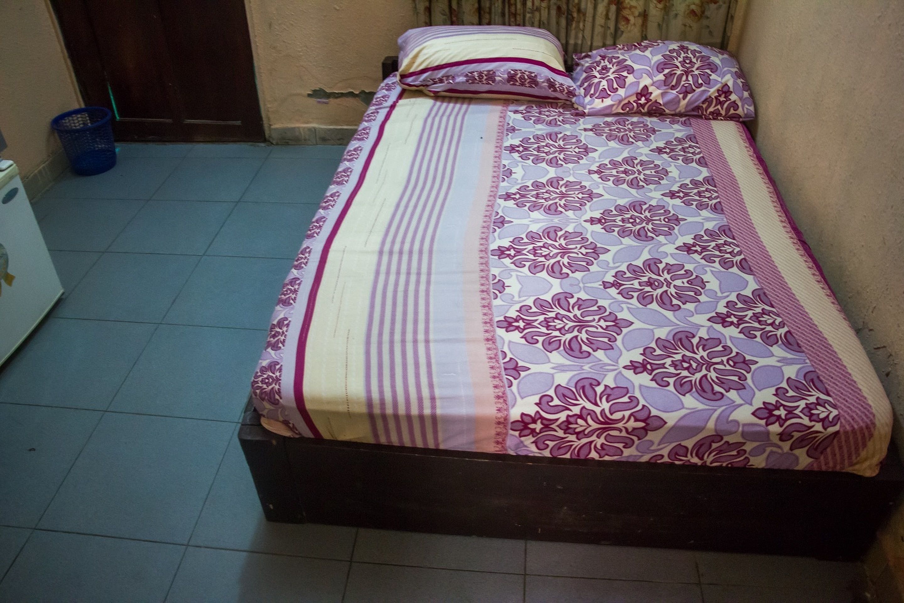 Un lit recouvert d’une couvre-lit à motifs floraux, agrémenté d’oreillers, un panier bleu et un appareil électroménager blanc, le tout dans une pièce dont le sol est carrelé.