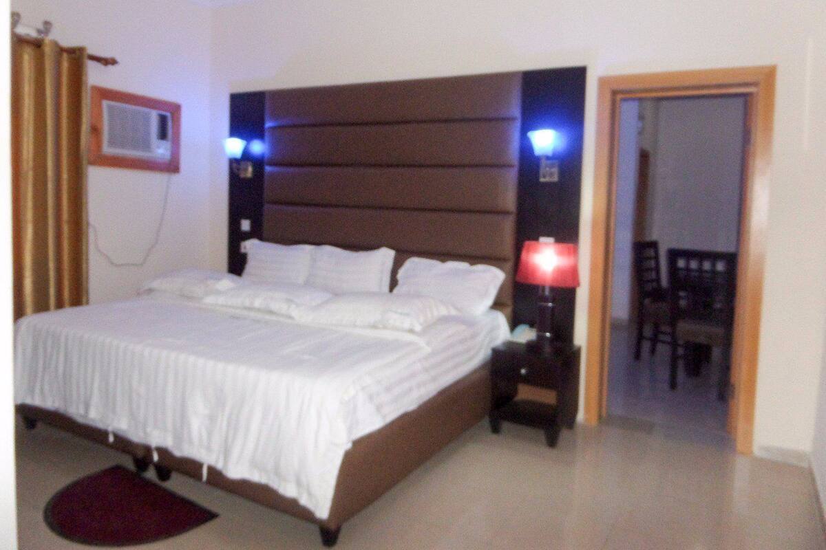 A hotel room with a bed, bedside lamps, a chair, and a door leading to another room.
