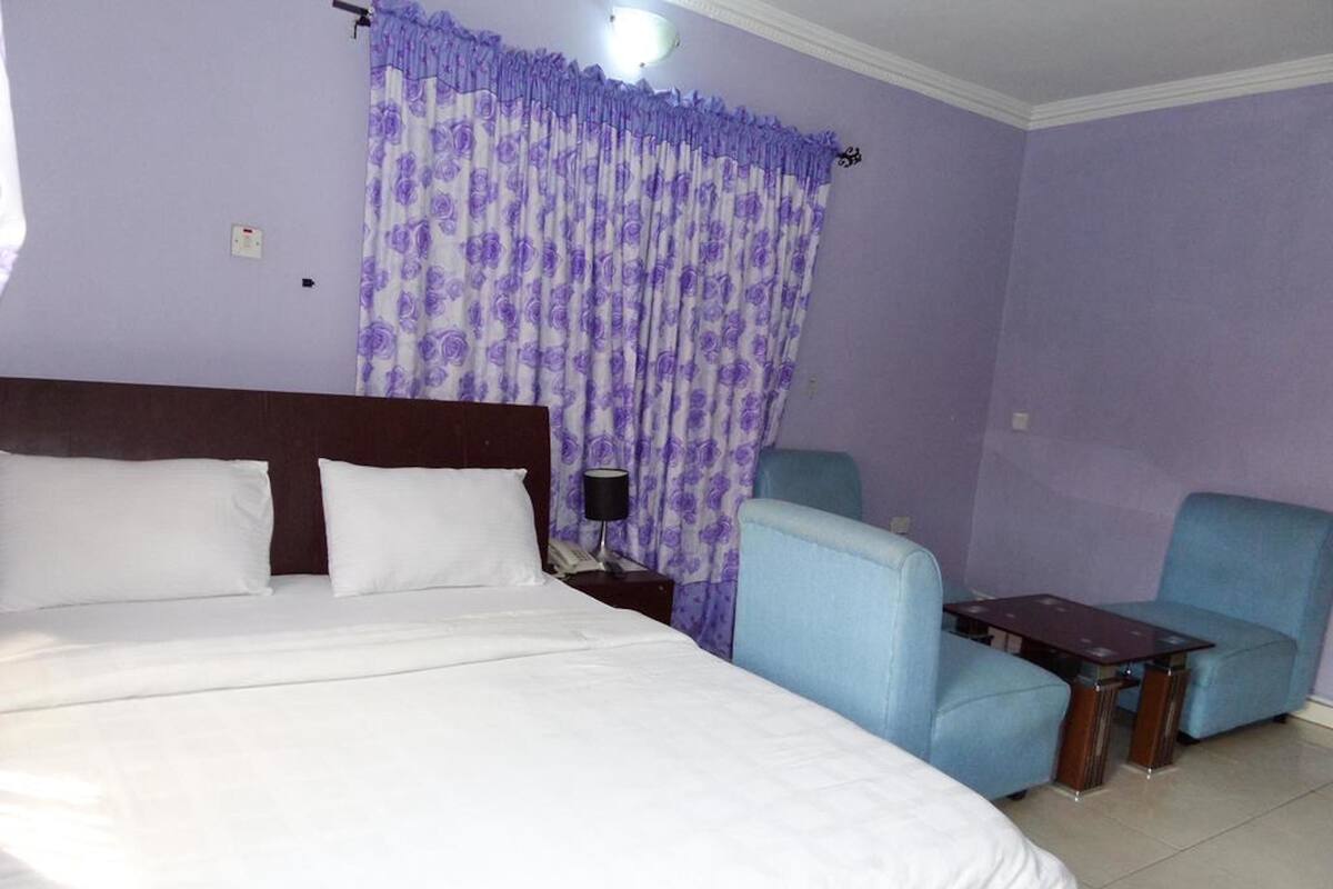 A hotel room with a large bed, two armchairs, a small table, and purple curtains.