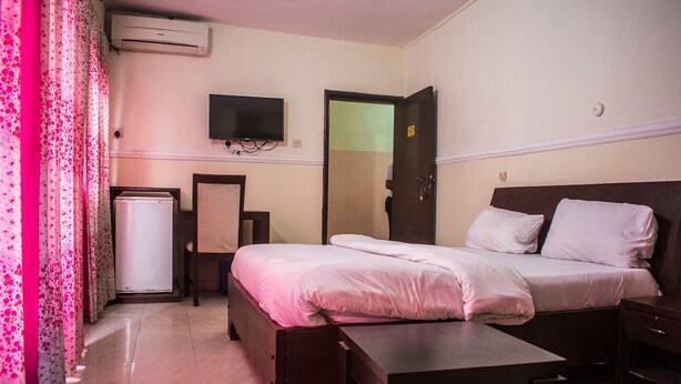 A hotel room with a bed, bedside table, a small desk, a television mounted on the wall, and a door. - Peerage Retreat and Resort (Lagos)
