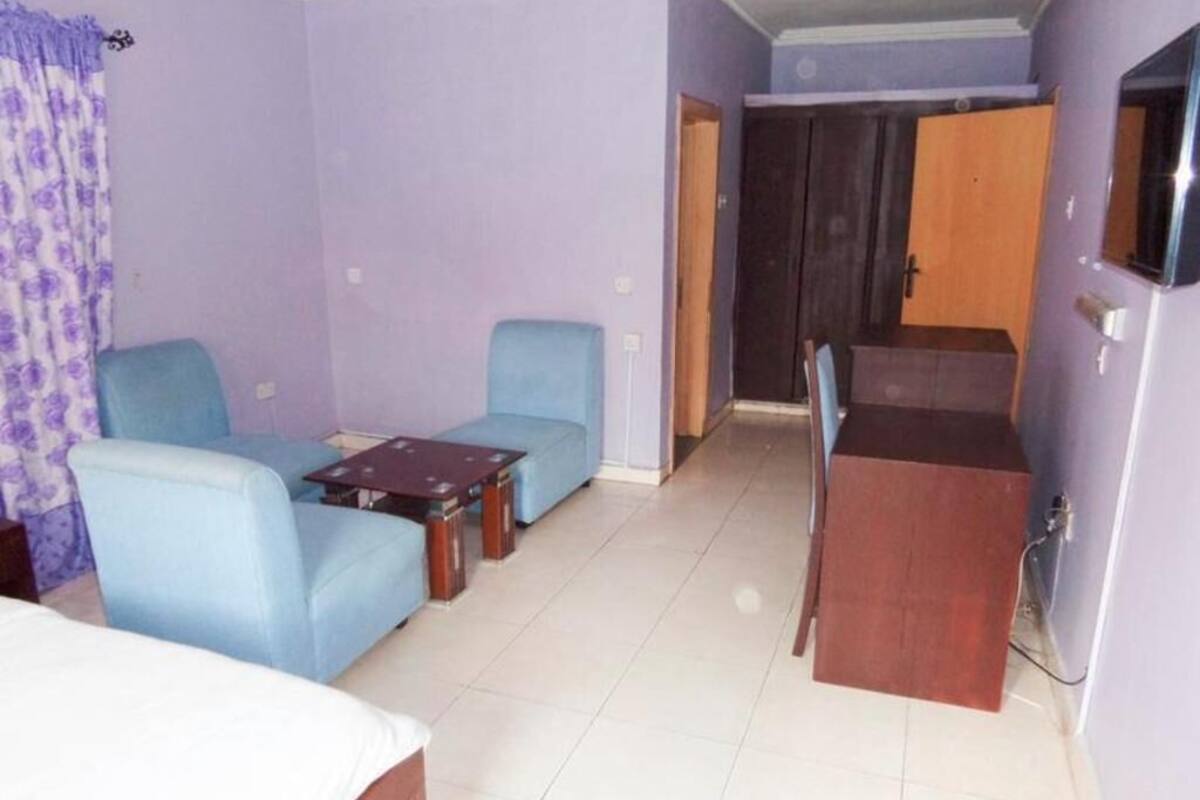 A room with a bed, two blue armchairs, a small table, a wooden cabinet, and a door leading to another room.