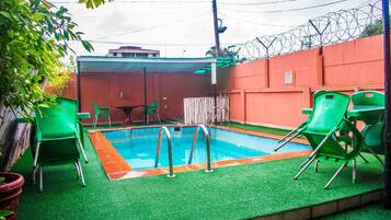 Une piscine avec des chaises vertes et un espace abrité, le tout entouré d’un mur élevé surmonté de barbelés.