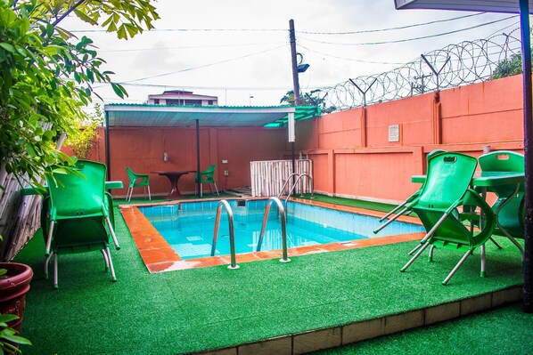 A swimming pool with green chairs and a covered area, surrounded by a high wall with barbed wire.
