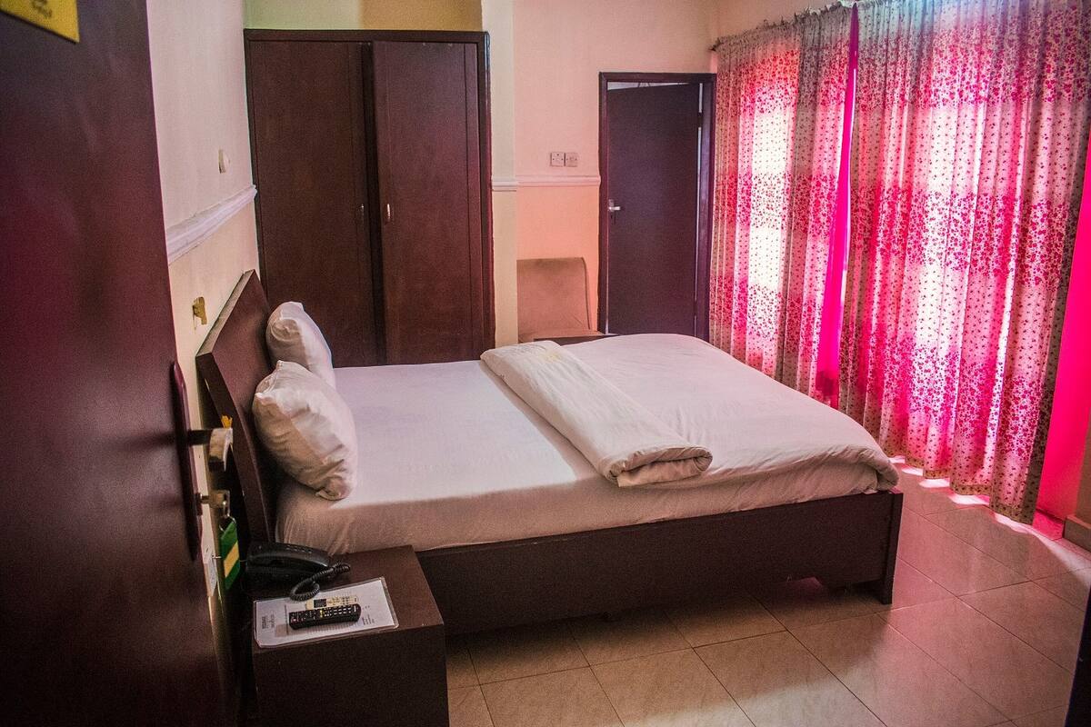 A single bed with a wooden frame, a bedside table with a telephone, and a curtain with a floral pattern.