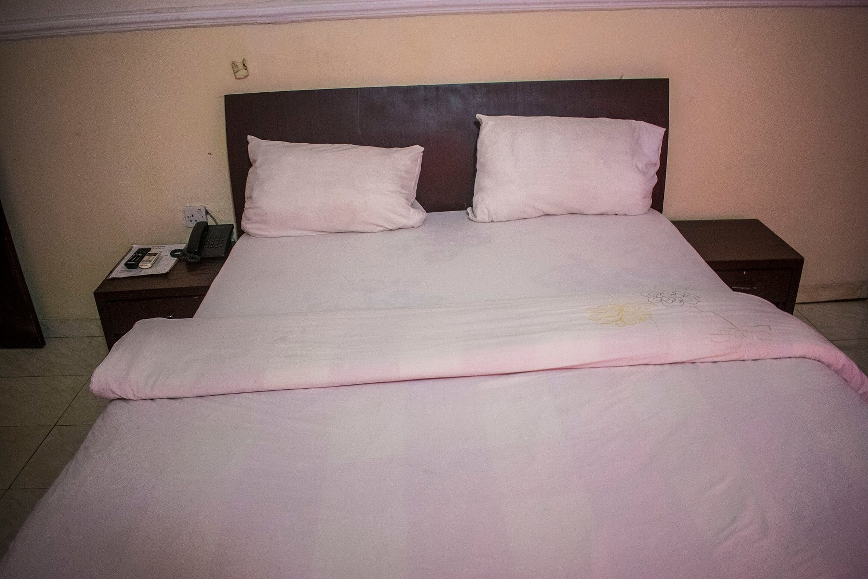 A neatly made bed with a white comforter and two pillows, a bedside table with a phone and a lamp, and a wall-mounted light switch.