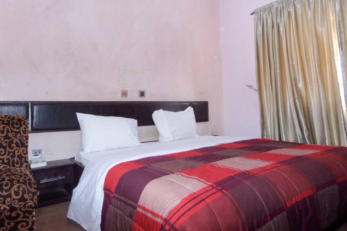 A hotel room with a large bed, a nightstand, a patterned armchair, and a window with curtains.