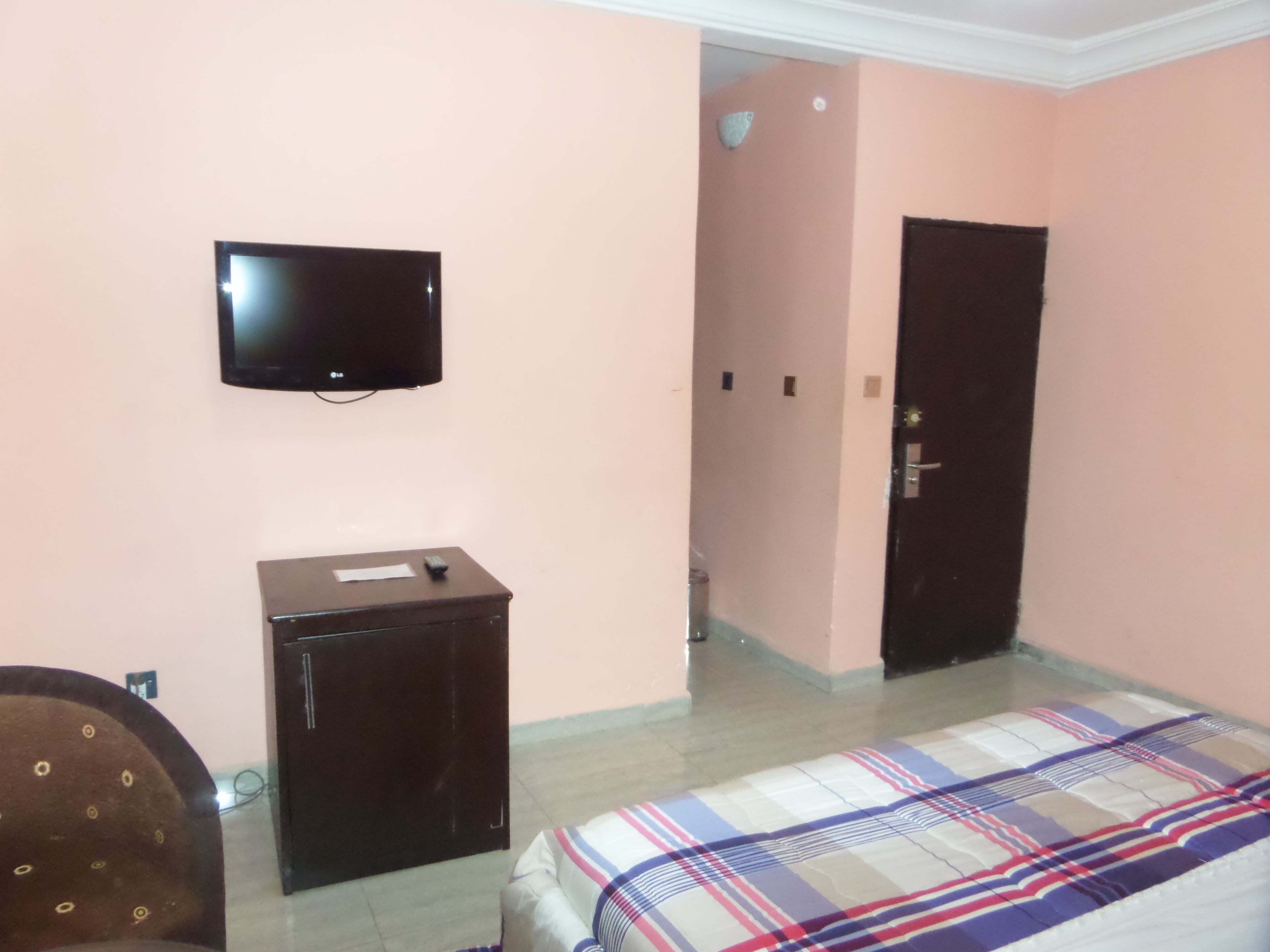 A bedroom with a bed, a television on the wall, a small cabinet, and a door leading to another room.
