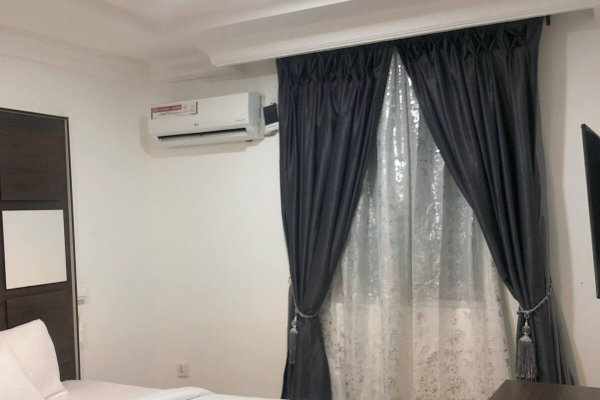 A hotel room with a bed, a desk, an air conditioning unit, and a window with curtains.