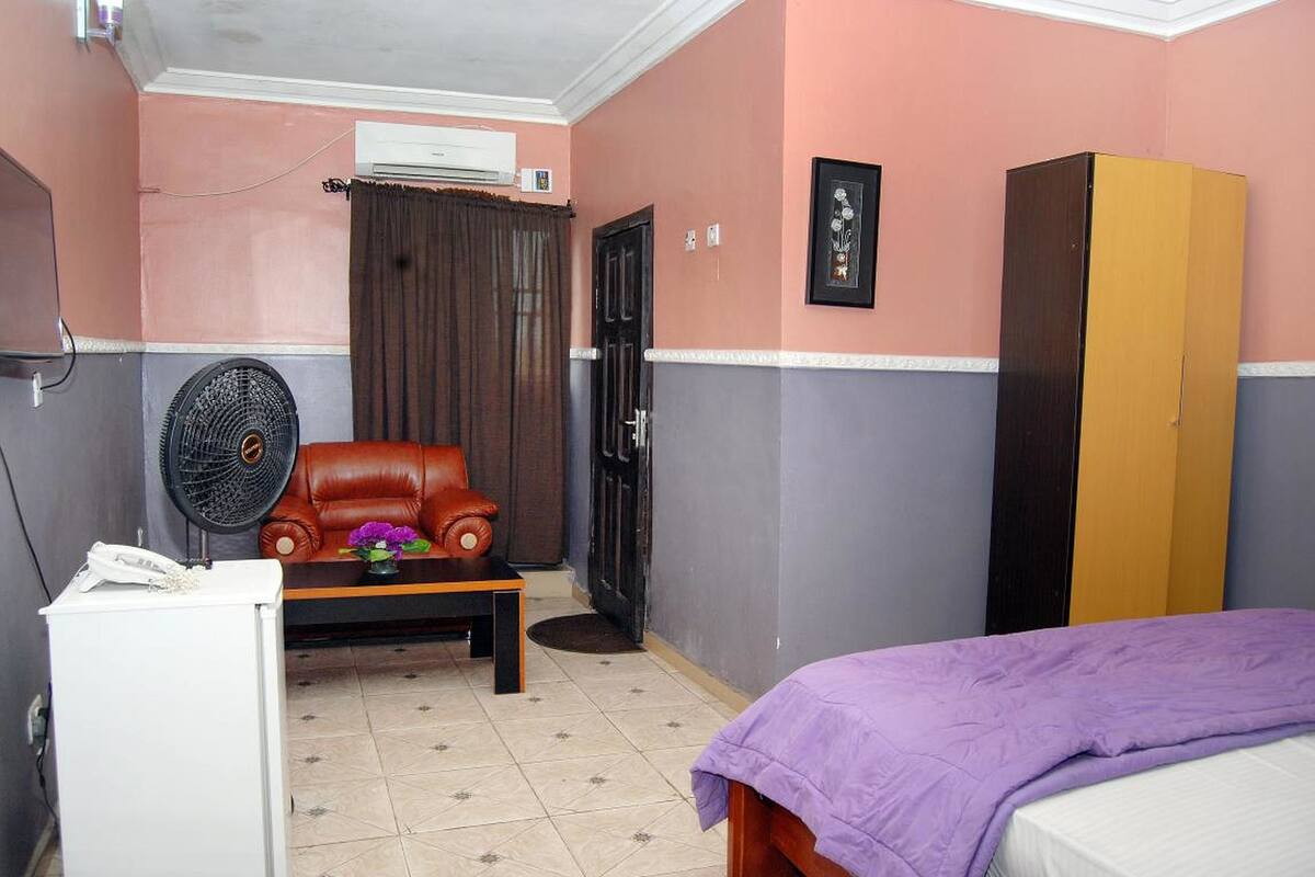 Una habitación con una cama, un escritorio con un ventilador, un sillón de cuero, un armario y una puerta.