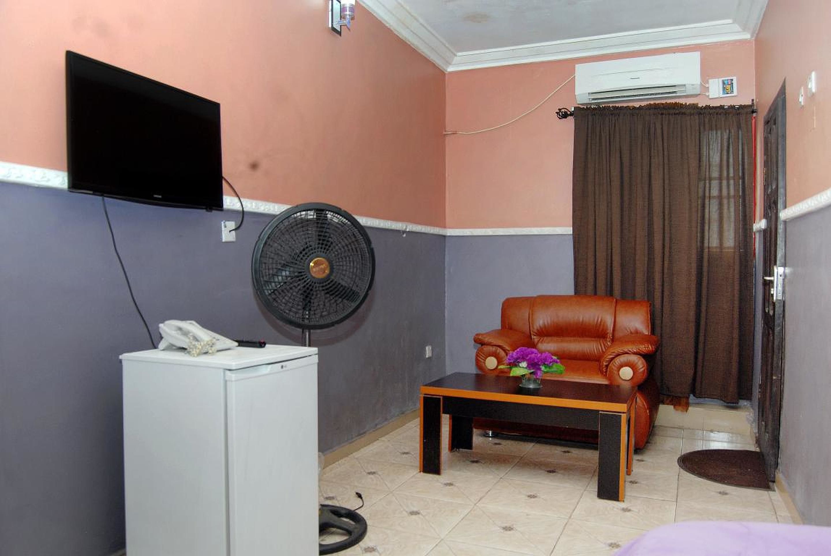 Una habitación compacta con una televisión de pantalla plana montada en la pared, un ventilador de pie, un sofá de cuero, una mesita con un florero y un refrigerador blanco.
