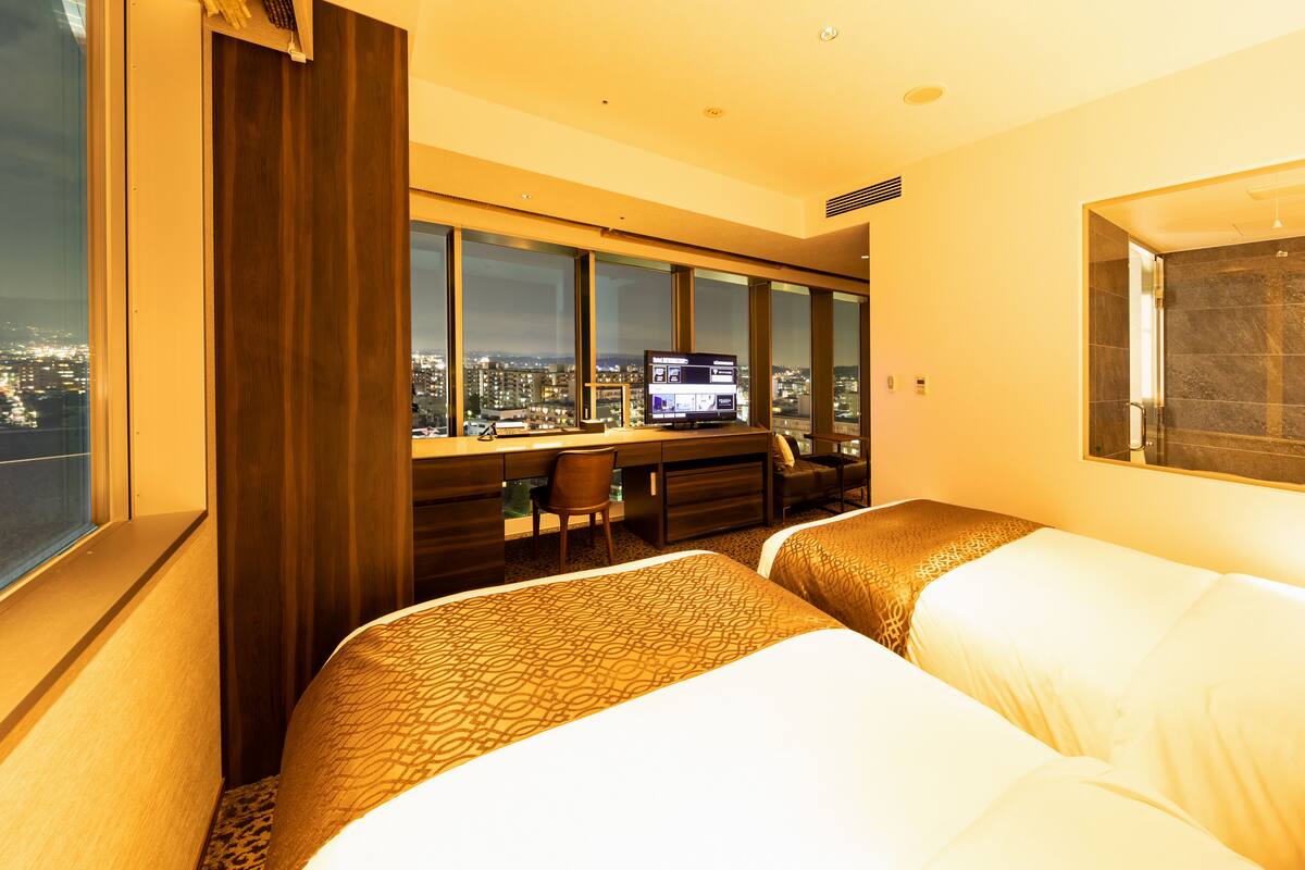 A hotel room with two beds, a desk, and a large window offering a city view.