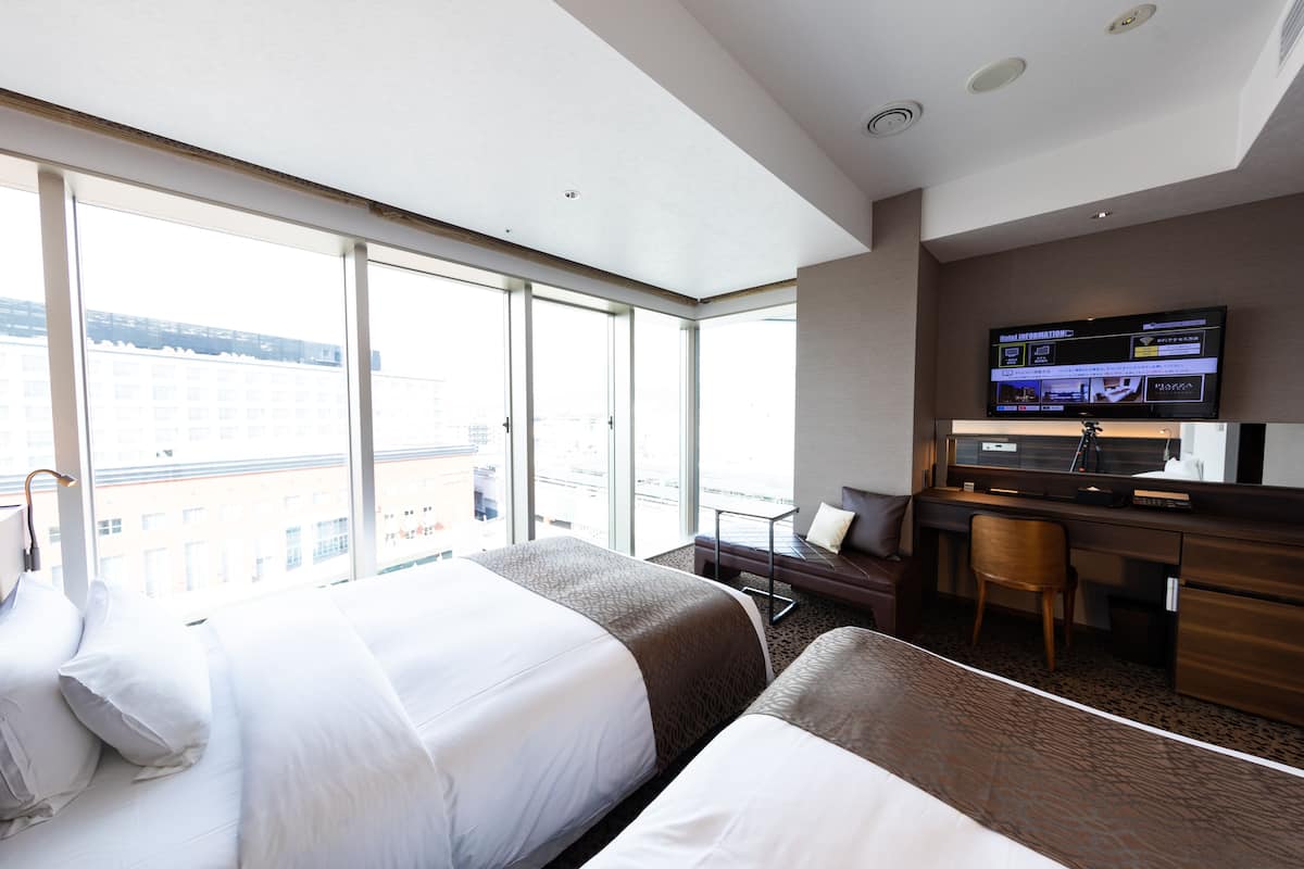 A hotel room with two beds, a desk, a chair, a TV, and large windows offering a city view.