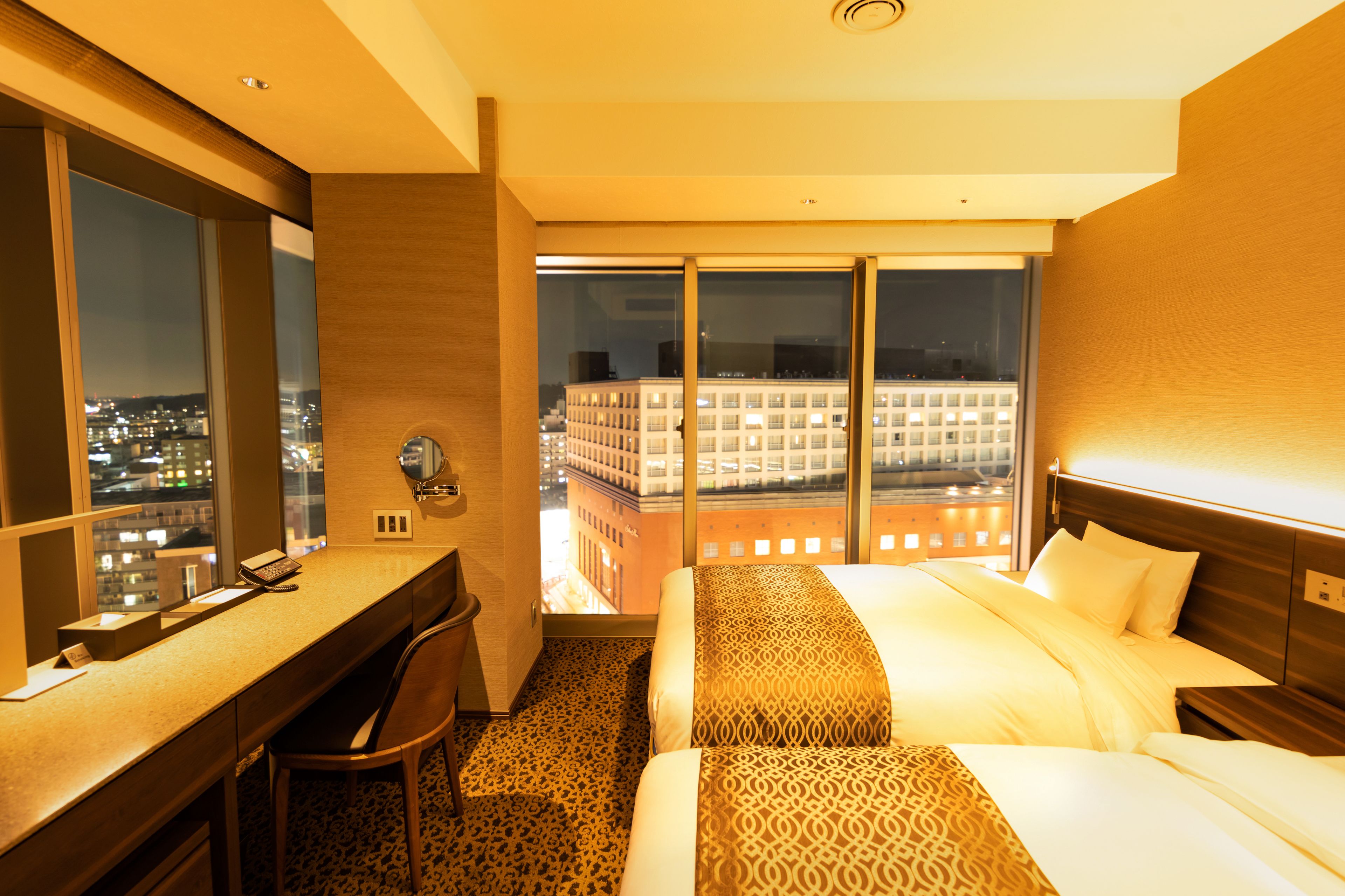 A hotel room with a desk, chair, bed, and a view of a cityscape through large windows.