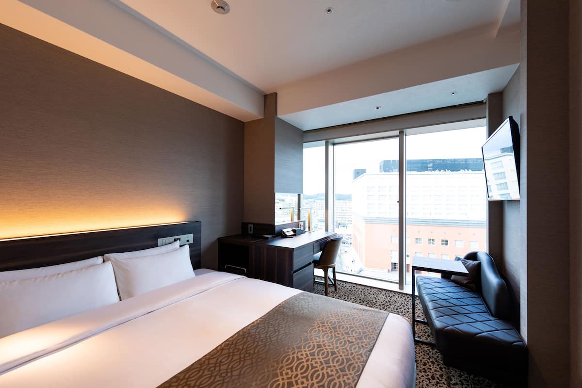 A hotel room with a large bed, a desk, a chair, and a sofa. There is a large window with a view of buildings outside.