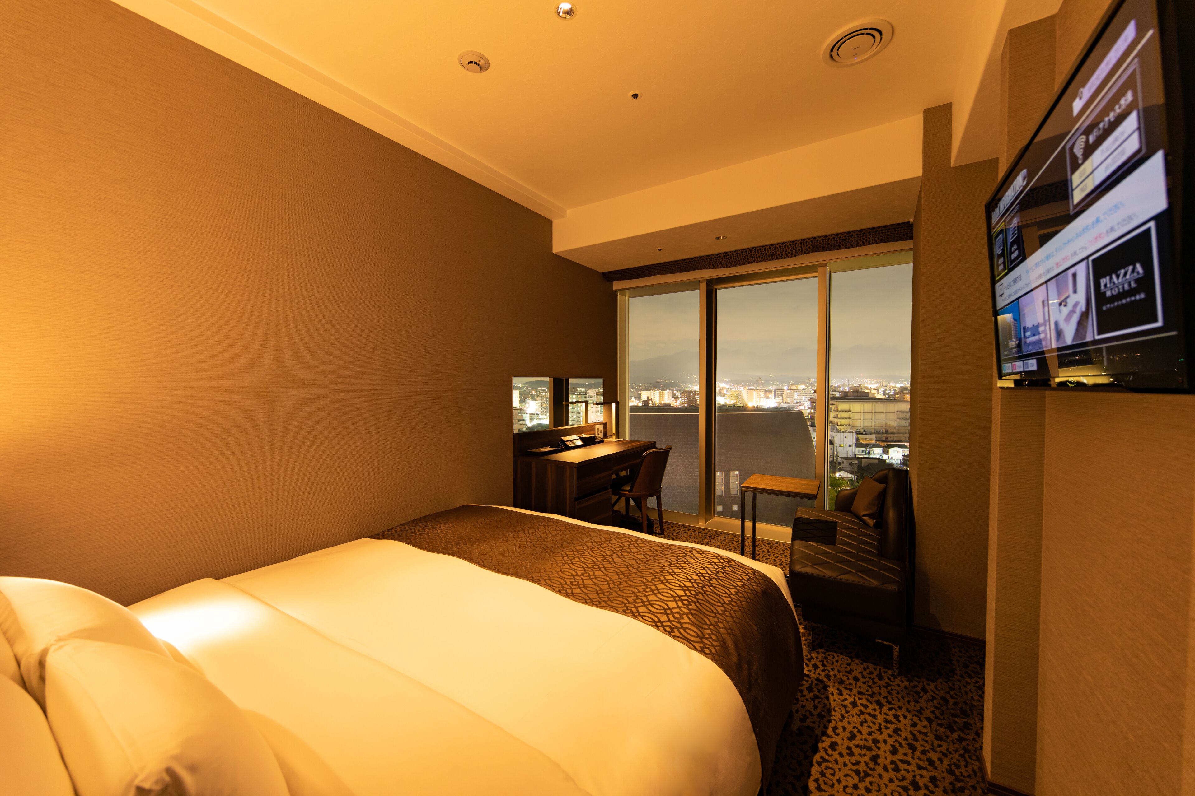 Habitación de hotel con una cama grande, un escritorio, una silla y un televisor. Se observa una vista de la ciudad desde la ventana.