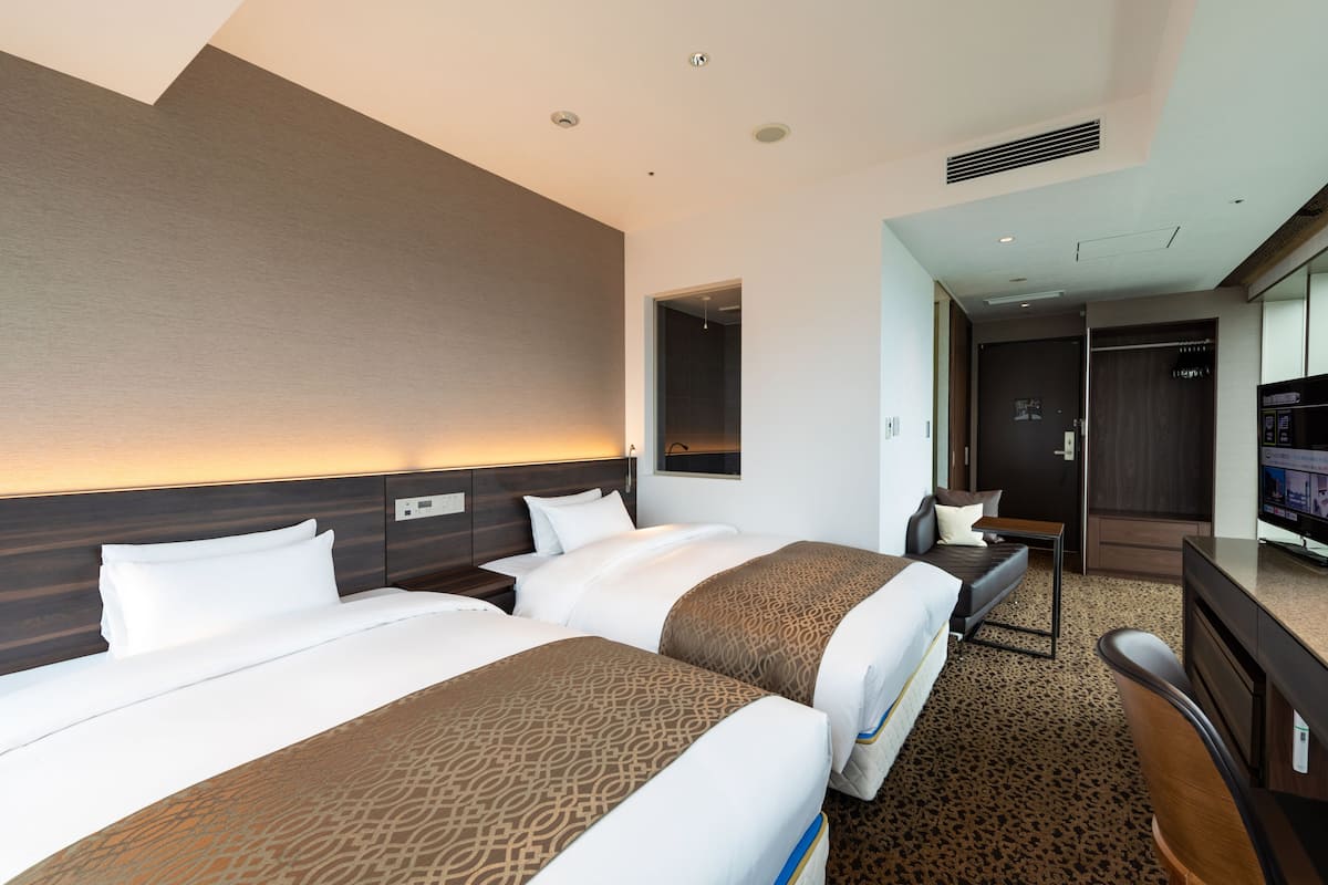 A hotel room with two beds, a desk, a chair, and a television.