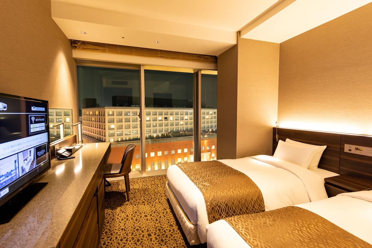 A hotel room with a large bed, a desk with a computer, and a view of a cityscape through large windows.