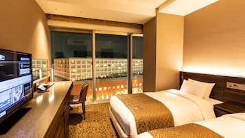 Habitación de hotel con una cama grande, un escritorio con computadora y vistas a un paisaje urbano a través de amplios ventanales.