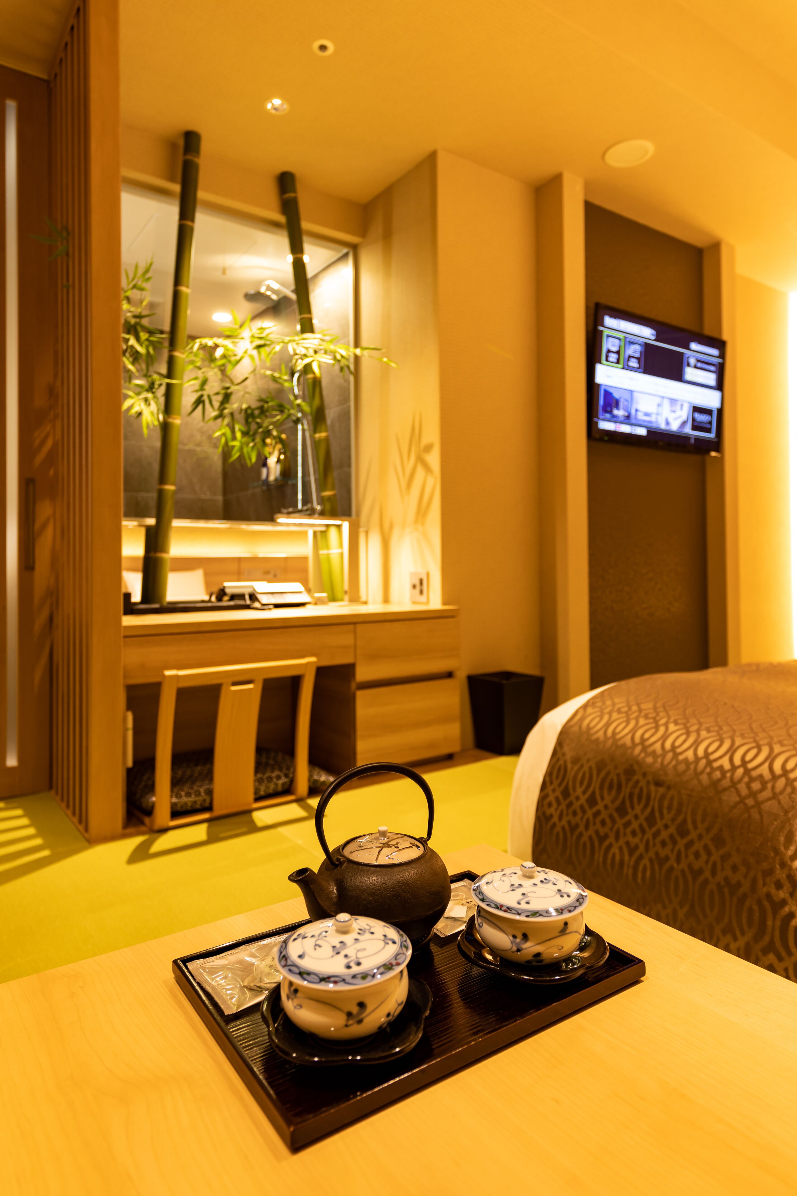 A hotel room with a desk, two teapots, a television, and a bed.