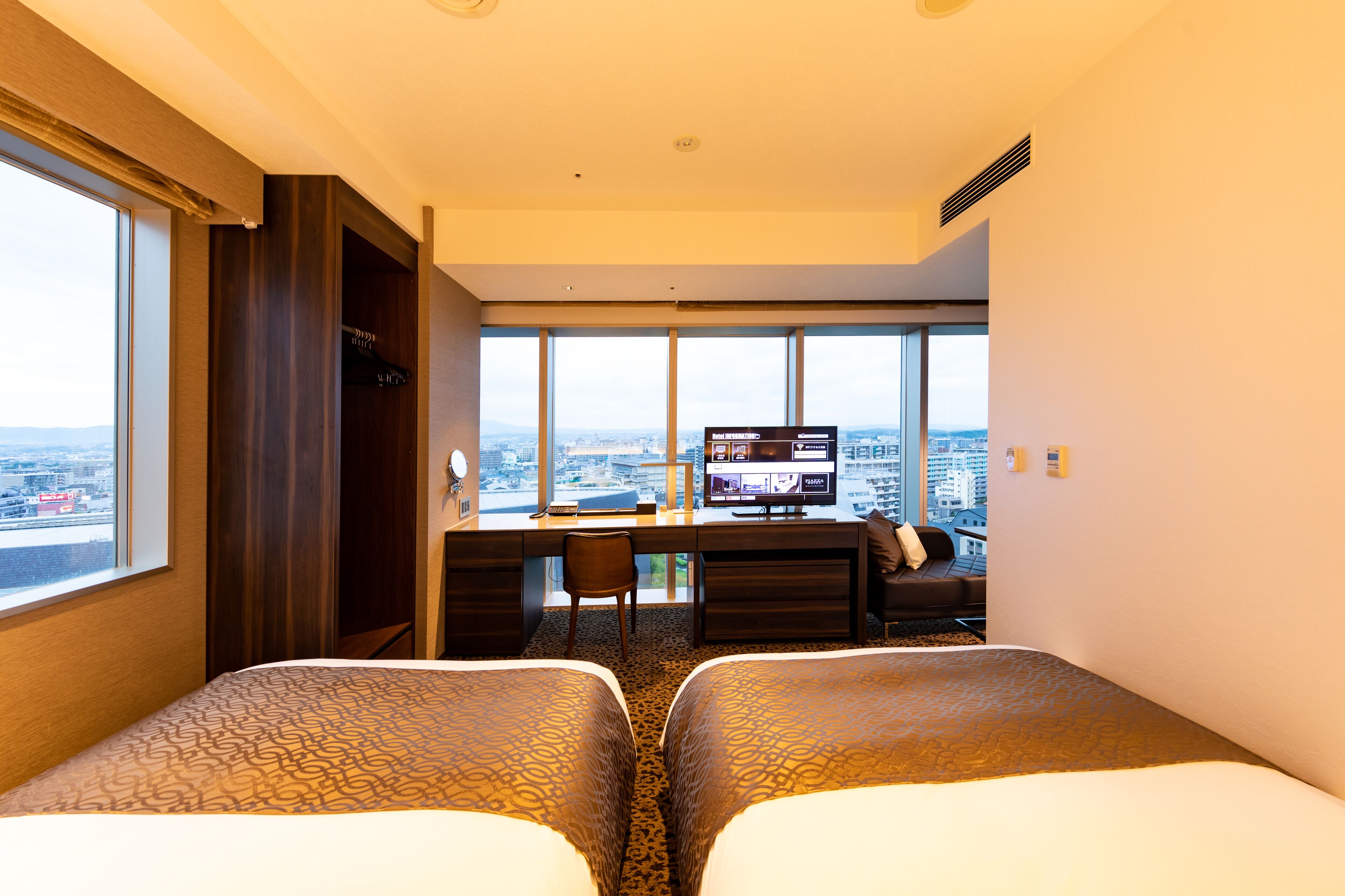 Een hotelkamer met twee bedden, een bureau met een televisie, en een groot raam met uitzicht op de stad.