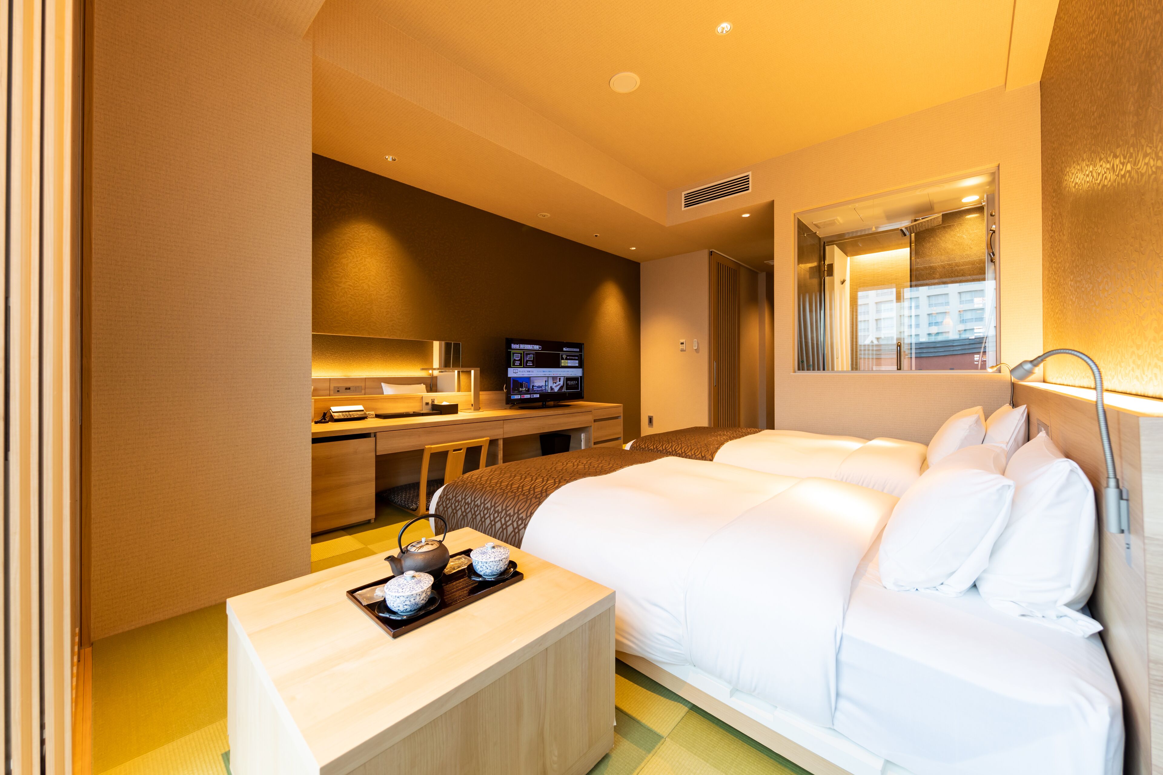 A hotel room with a bed, a desk with a television, and a small table with teapots.