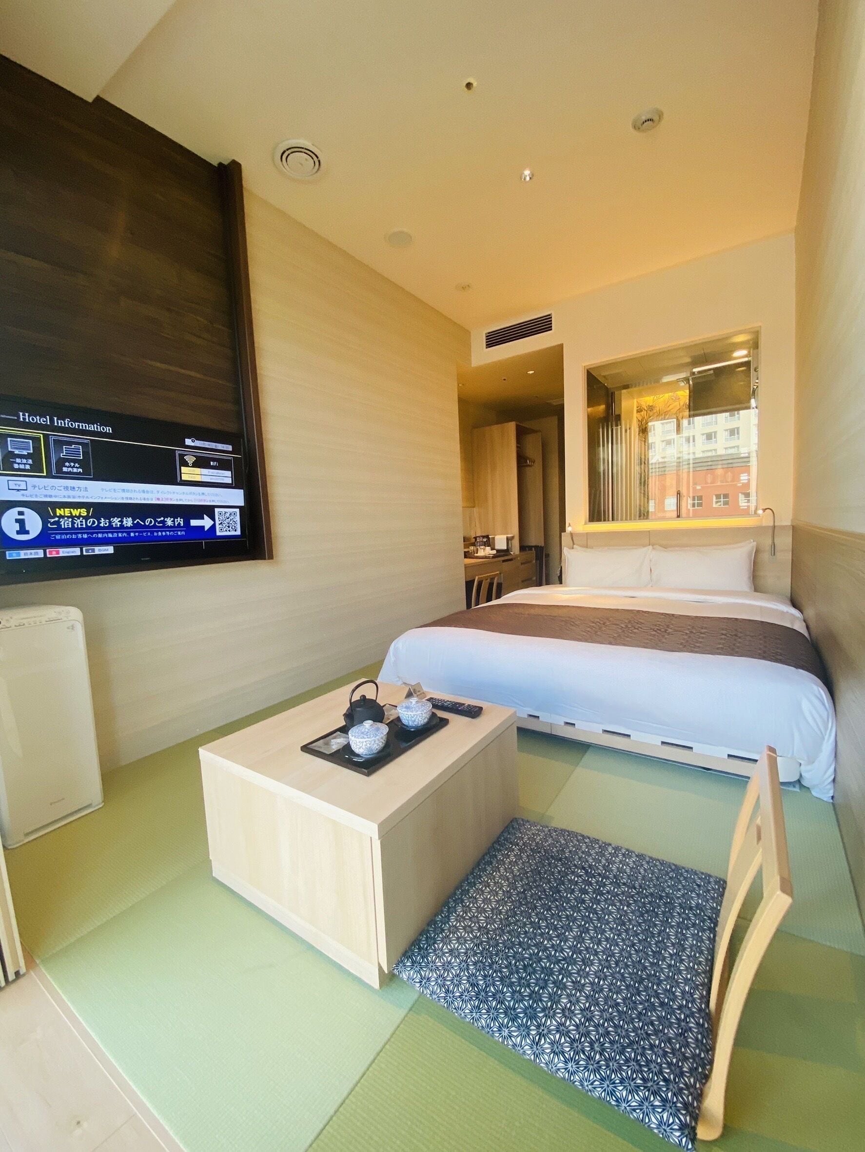 A hotel room with a bed, a desk with a kettle and cups, a chair, and a TV displaying hotel information.