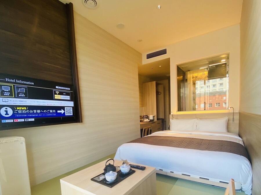 A hotel room with a bed, a desk with a kettle and cups, a chair, and a TV displaying hotel information.