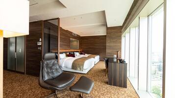 Ein modernes Hotelzimmer mit einem großen Bett, einer Sitzecke, einem Schreibtisch und Blick auf die Stadt durch große Fenster.